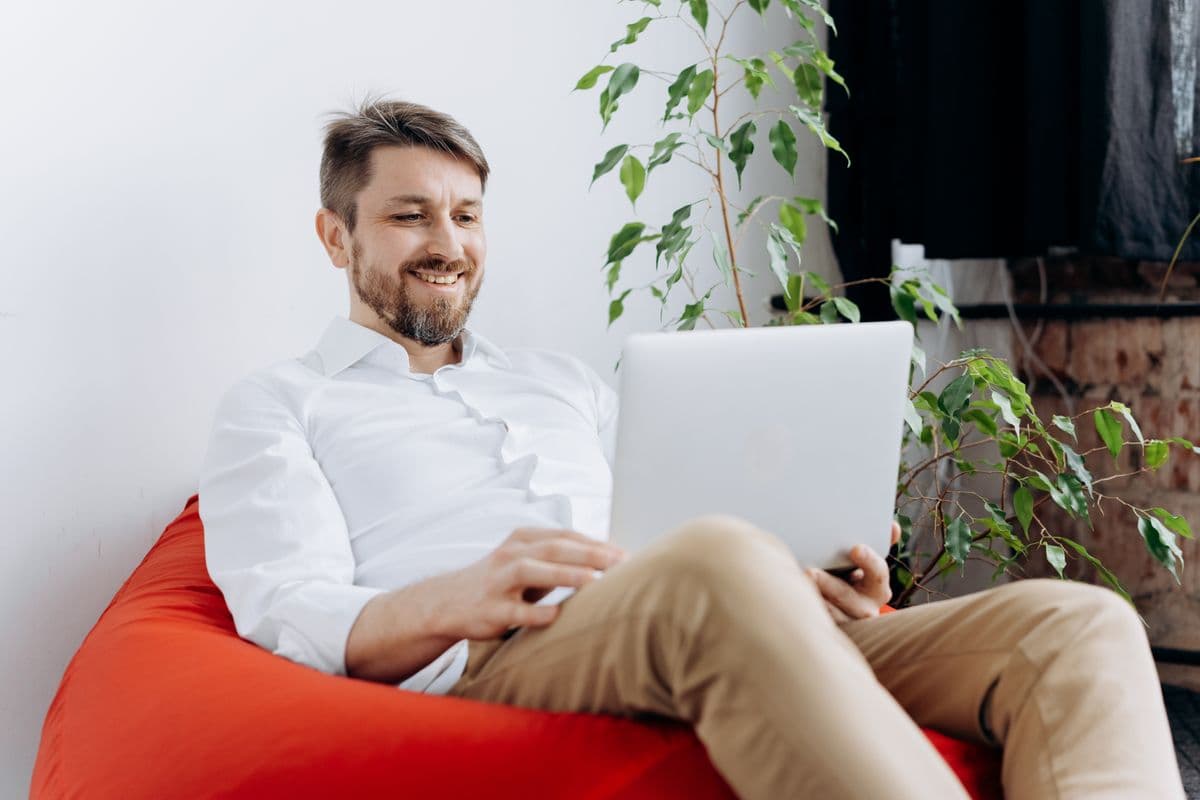 A smiling parent using a laptop at home beside a large leafy houseplant